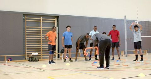 Diverse male athletes training with hurdles and volleyball in indoor gym drill session