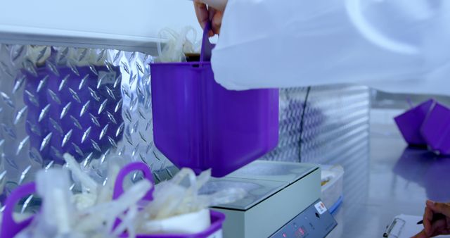 Image shows a close-up of a laboratory worker using a weighing scale. A purple container is being placed on the digital scale. The background has a metallic texture indicating a lab environment. This image can be used to represent scientific research, precise measurement, laboratory activities, or technological advancements in healthcare. Ideal for educational materials, professional presentations, and marketing for scientific products or laboratories.