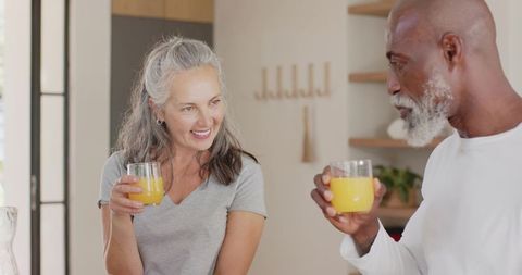 Mature diverse couple smiling and drinking orange juice in sunlit minimalist kitchen