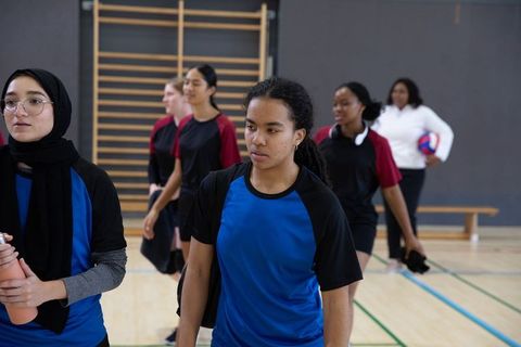 Diverse girls volleyball team walking in gymnasium, focused teen leader in blue jersey