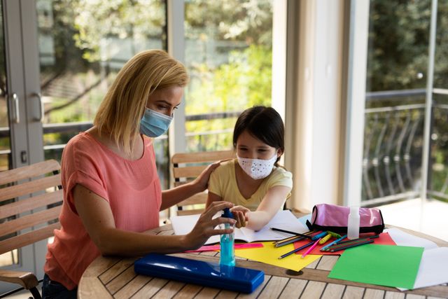 Caucasian mother and daughter practicing good hygiene by using hand sanitizer while studying at home. Both are wearing masks, emphasizing safety and health during the COVID-19 pandemic. This image can be used for educational materials, health and safety campaigns, or articles about homeschooling and pandemic precautions.