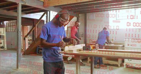 Carpenter marking wood with pencil wearing safety goggles and earmuffs in workshop