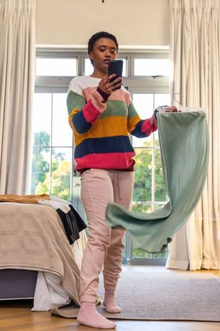 African american woman holding phone and dress, choosing casual outfit in sunlit bedroom