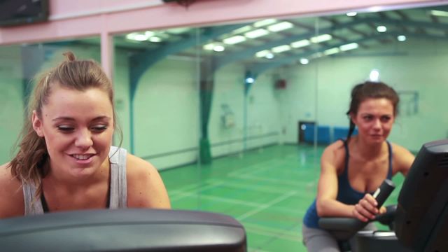 Two women riding exercise bikes together in a gym while chatting creates a vibrant atmosphere of camaraderie and health. This can be used for promoting gym memberships, fitness classes, or health and wellness campaigns, underlining benefits of a social workout setting.