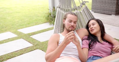Diverse couple relaxing in garden hammock with smartphone