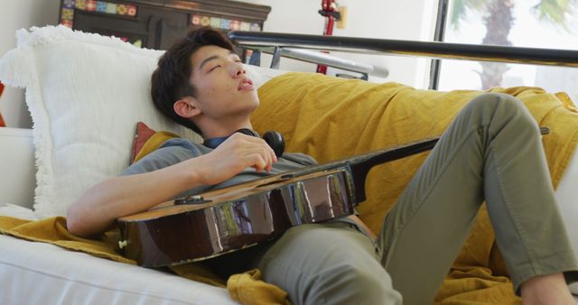 Asian male teenager with headphones playing guitar and lying in living room. spending time alone at home.