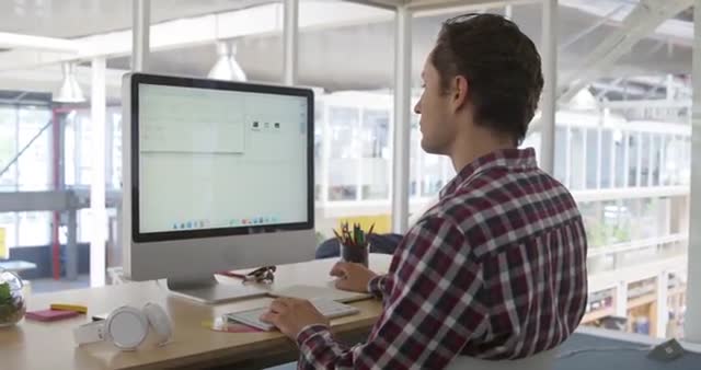 Young businessman focusing on work at computer station in open modern office, suggesting innovation and productivity. Ideal for use in corporate, technology, and workplace-related content.