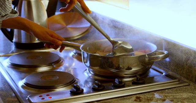 Person stirs pasta in a pot on a contemporary stove. Steam rises from the pot, indicating that the pasta is cooking. Bright kitchen lighting adds a clean and modern atmosphere. Ideal for showing home cooking, kitchen appliances, recipe demonstrations, culinary blogs, or advertisements for cooking products.