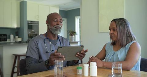 Home nurse consulting patient about medication with tablet and pill bottles at kitchen table