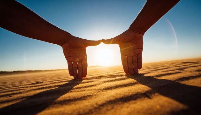 Hands framing sun over rippling sand dunes during golden hour creating geometric glow