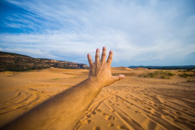 Hand is stretched out towards expansive desert with sand dunes, under clear blue sky. Could be used for concepts like adventure, exploration, journey, and longing. Great for travel blogs, outdoor adventure marketing, or motivational themes.