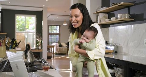 Mother Holding Baby Daughter in Kitchen Watching Laptop Pot Kettle Mug