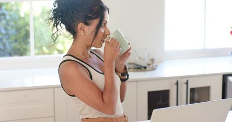 Woman sipping coffee and checking laptop at sunlit modern kitchen island for home office