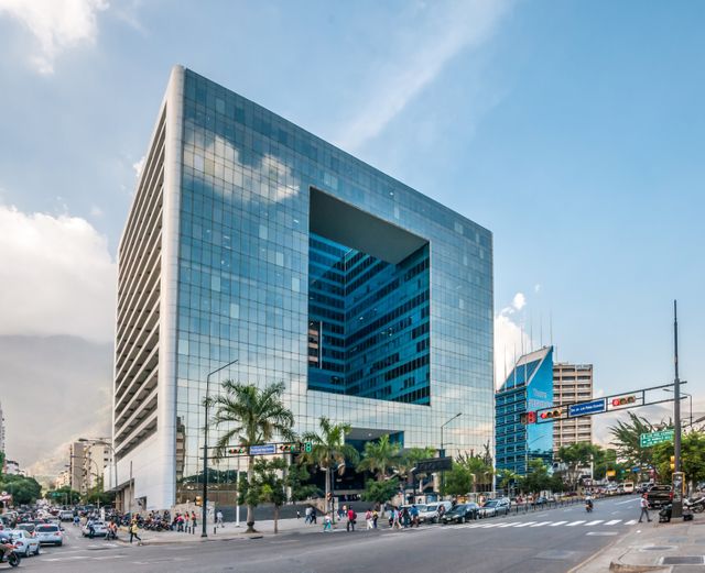 Glass office building with an unusual design located in busy urban area featuring palm trees and city life with automobiles and pedestrians. Suitable for businesses, real estate promotions, corporate layouts, architectural magazines, urban lifestyle blogs.