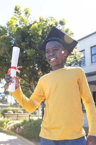 African american boy celebrating graduation outdoors wearing mortarboard holding diploma
