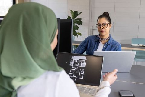 Diverse women reviewing architectural floorplan on laptop in collaborative office workspace