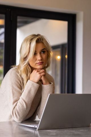 Blonde woman working on laptop at home, gazing pensively, modern minimalist kitchen
