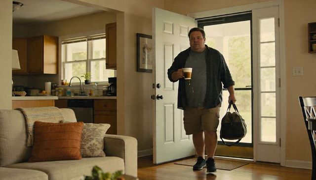 Man entering home carrying duffel while spilling coffee casual plaid shirt khaki shorts