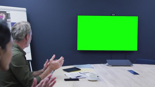 Diverse business professionals sitting in conference room applauding during video call with green screen displayed on television. Useful for illustrating themes of remote work, business meetings, diversity in the workplace, and team collaboration.