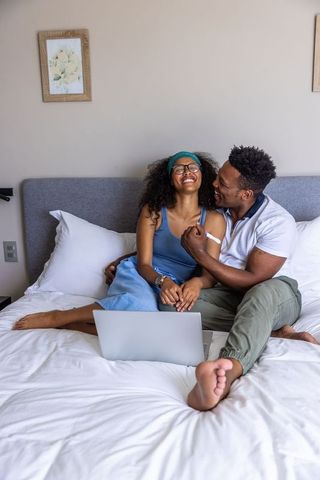 Laughing romantic diverse couple relaxing on bed with laptop embracing in cozy bedroom