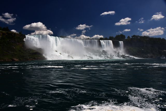 Niagara Falls, depicted here cascading dramatically under a vibrant blue sky with scattered clouds, showcases its immense power. Perfect for use in travel magazines, promotional tourism materials, environmental awareness campaigns, and nature documentaries highlighting North American landmarks.