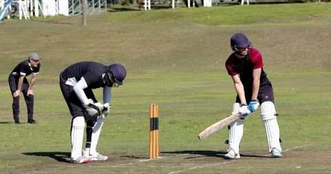 Cricket batsman batting at crease while keeper adjusting gloves behind stumps on grass