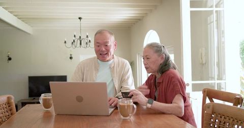 Asian senior couple using laptop and holding credit card at home dining table with coffee