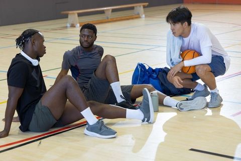 Three multiracial athletes resting and talking on indoor basketball court after practice