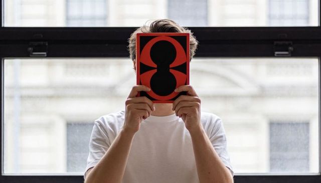 Anonymous reader hiding face behind red graphic book at urban window, conceptual portrait