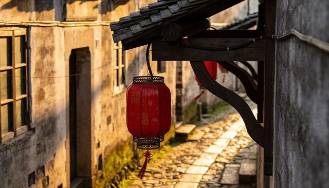 Red chinese lantern hanging under tiled eave over sunlit cobblestone alleyway at warm dusk