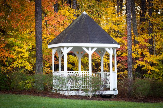 White gazebo stands amidst vibrant autumn foliage in tranquil park. Ideal for depicting serenity, nature, fall season, and picturesque outdoor settings. Use in articles related to autumn recreational activities, seasonal photography, or relaxing nature scenes.