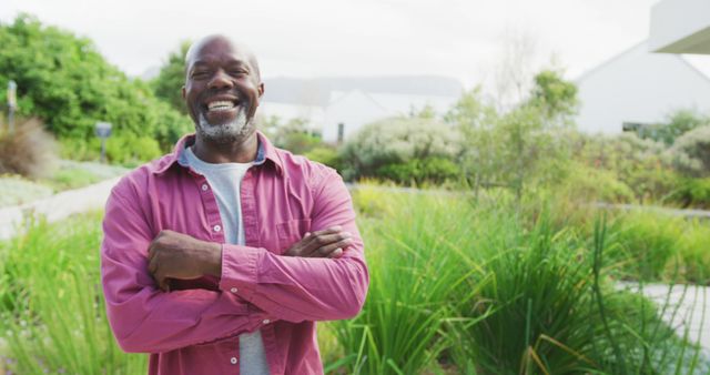 This image depicts a mature man smiling with arms crossed while standing outdoors. The background features greenery and hints of residential houses, suggesting a relaxing environmental setting. This could be used to represent themes of relaxation, senior lifestyle, happiness, self-confidence, and outdoor enjoyment.