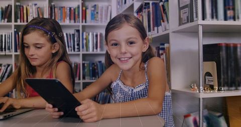Girls collaborating with tablet and laptop in library smiling while learning technology