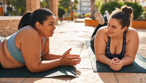Plus-size women chatting on yoga mats on sunlit terrace, exercising and relaxing together
