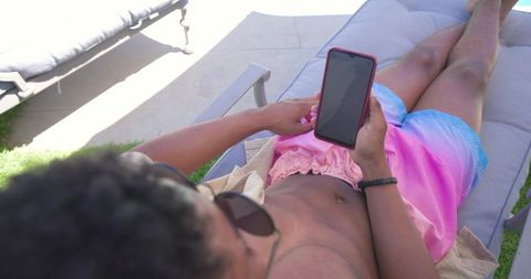 Reclining man holding smartphone on poolside lounge chair wearing pink blue swim shorts