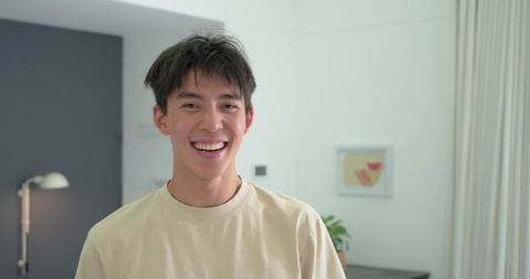 Asian man smiling and standing in modern minimalist living room wearing casual beige tee