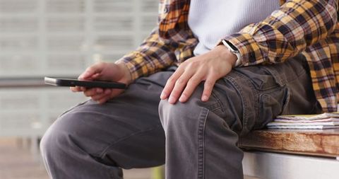Asian man sitting on wooden bench wearing plaid shirt and cargo pants holding smartphone