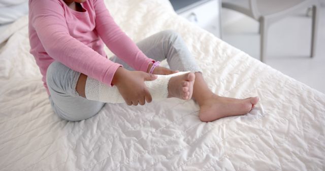 Child wrapping foot with bandage while sitting on bed. Suitable for health and wellness articles, injury prevention guides, physical therapy resources, and family care blogs.