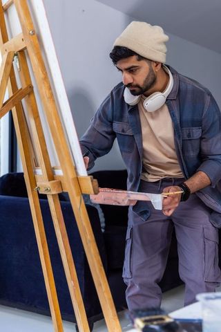Young man painting on canvas in home studio wearing beanie and headphones