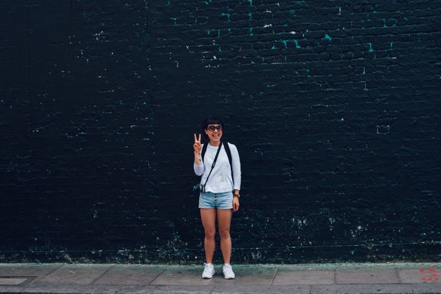 Cheerful Asian woman standing in front of a dark urban wall, making a peace sign with her fingers. She is wearing casual summer clothing, including shorts and a white shirt, capturing a moment of happiness in a cityscape environment. Perfect for use in lifestyle blogs, websites addressing urban living, fashion brands promoting casual wear, or social media posts emphasizing positivity and youth culture.
