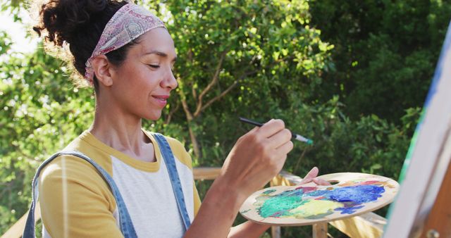 Woman in casual attire stands outdoors surrounded by trees. She holds a palette filled with various colors and paints on a canvas. Ideal for showcasing creativity, activities involving art, and the joy of painting in natural settings.