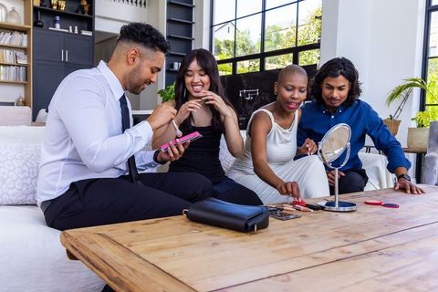 Diverse friends sharing makeup, trying looks and swapping phone in bright living room