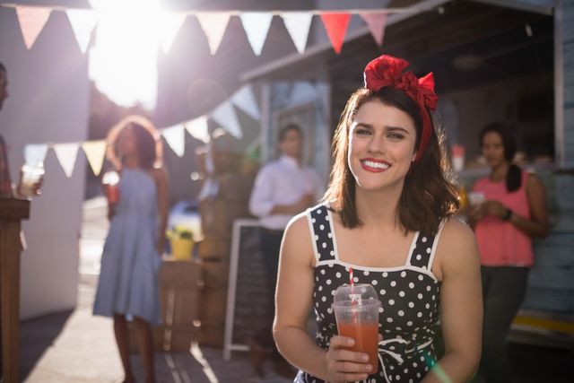 Smiling woman holding a glass of juice at an outdoor event with bunting and people in the background. Ideal for use in advertisements, social media posts, and articles about summer events, outdoor gatherings, and festive celebrations.