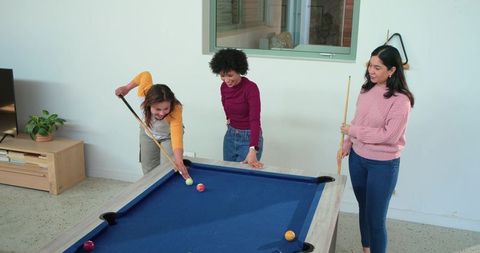 Diverse female friends playing pool, leaning around blue-felt table and laughing in home lounge