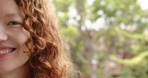 Red-haired woman smiling closeup showing curly hair and freckles among sunlit green bokeh
