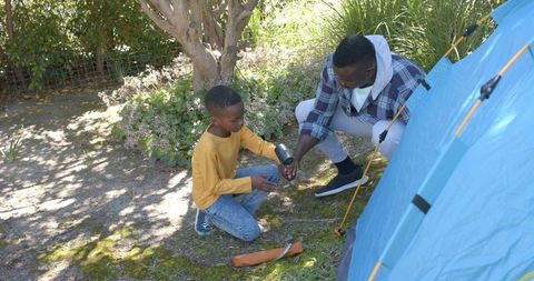 Father and son pitching backyard tent and sharing metal cup while camping and bonding