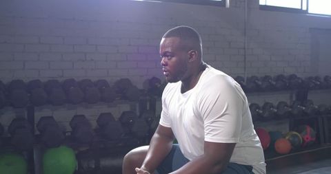 African American man resting on gym bench wearing white tee and shorts near dumbbells