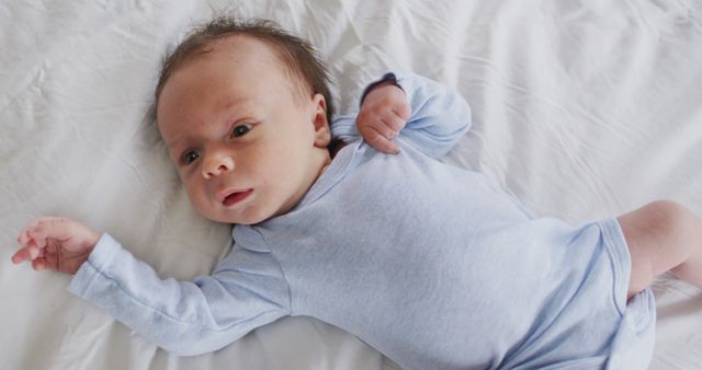 Newborn baby wearing a light blue onesie lying on a white bed, looking up with arms slightly raised. Ideal for use in parenting blogs, infant care articles, baby product advertisements, and nursery room decor.