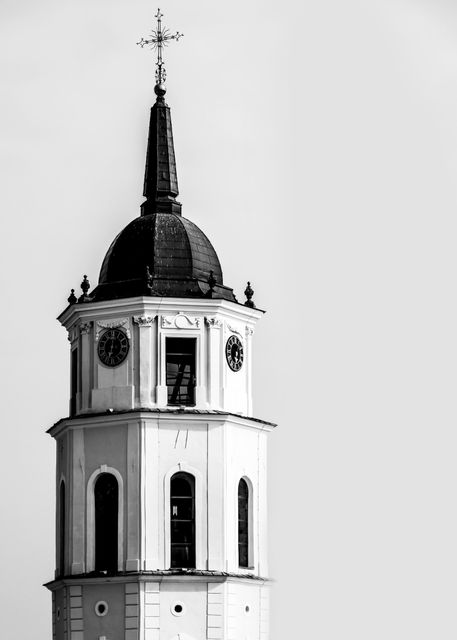 Monochrome photograph depicting a historic church tower topped with a clock and intricate steeple against a clear sky. Suitable for use in projects related to historic sites, architecture, religious themes, or travel. Ideal for websites, brochures, or educational materials on history and architecture.