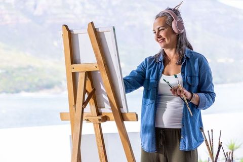 Senior woman painting on coastal terrace wearing pink headphones holding brushes